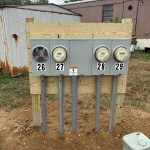 Four electric meters mounted on a wooden panel, with a warning sign, near a mobile home and storage shed.
