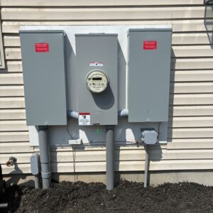Utility meter and electrical panels mounted on a house exterior, featuring warning labels and connected conduits.