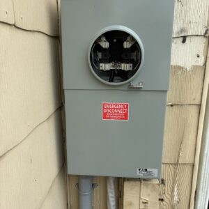 Gray electrical meter box mounted on a wall, featuring an emergency disconnect warning label.